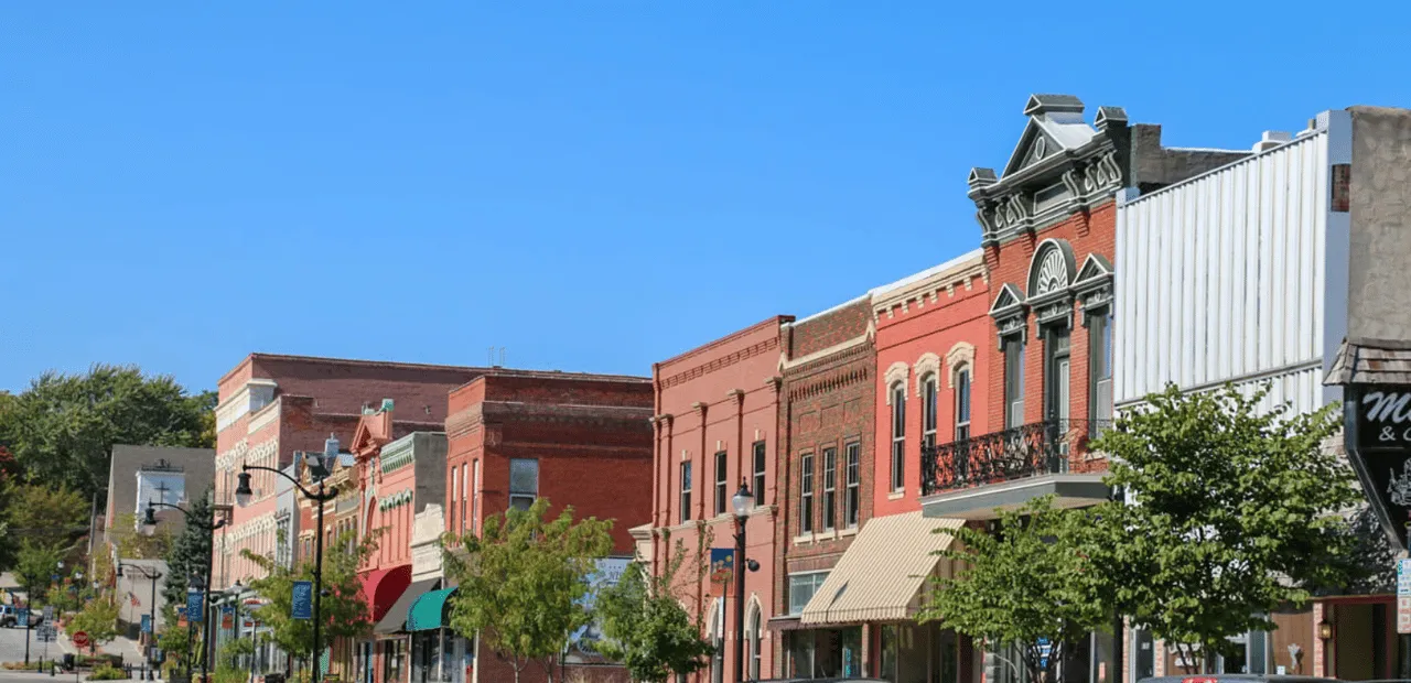 Historic brick storefronts and leafy trees line a sunny neighborhood street.
