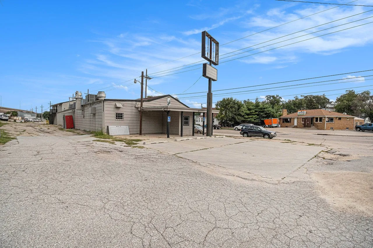 Commercial building with a covered entrance and cracked asphalt parking area beneath a tall roadside sign frame.