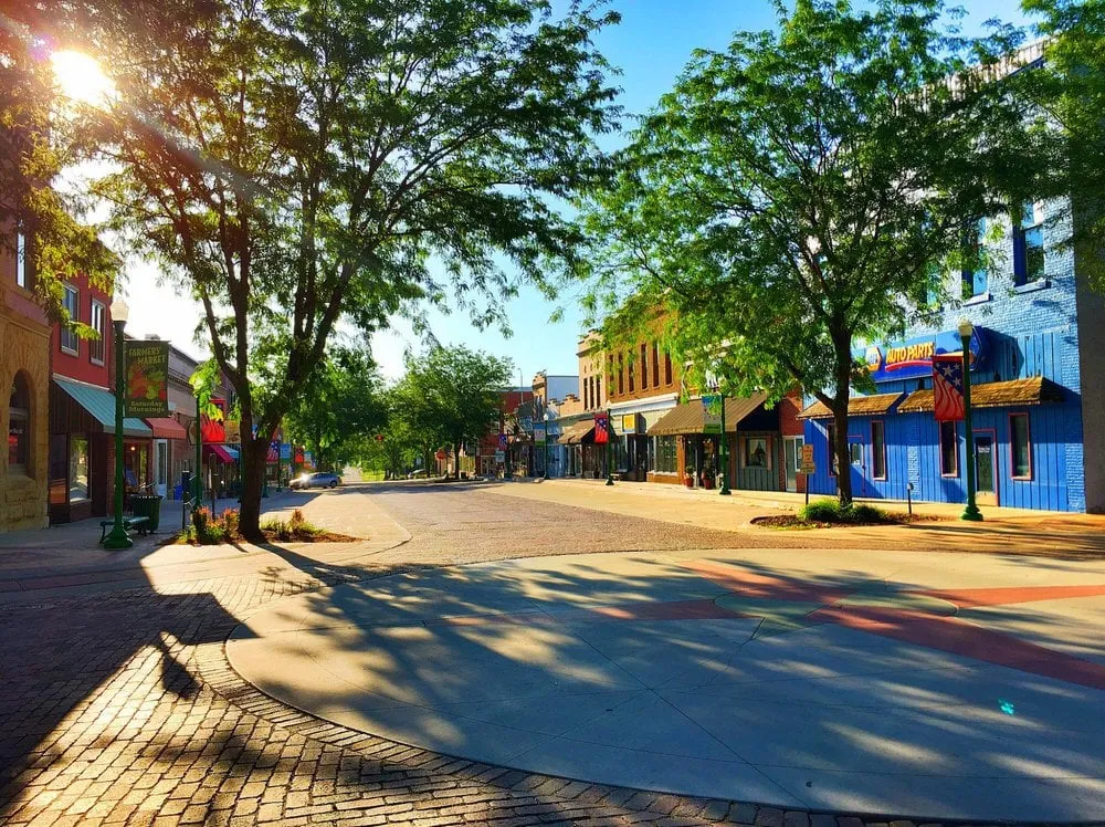Tree-lined neighborhood street with colorful storefronts and a farmers market sign.