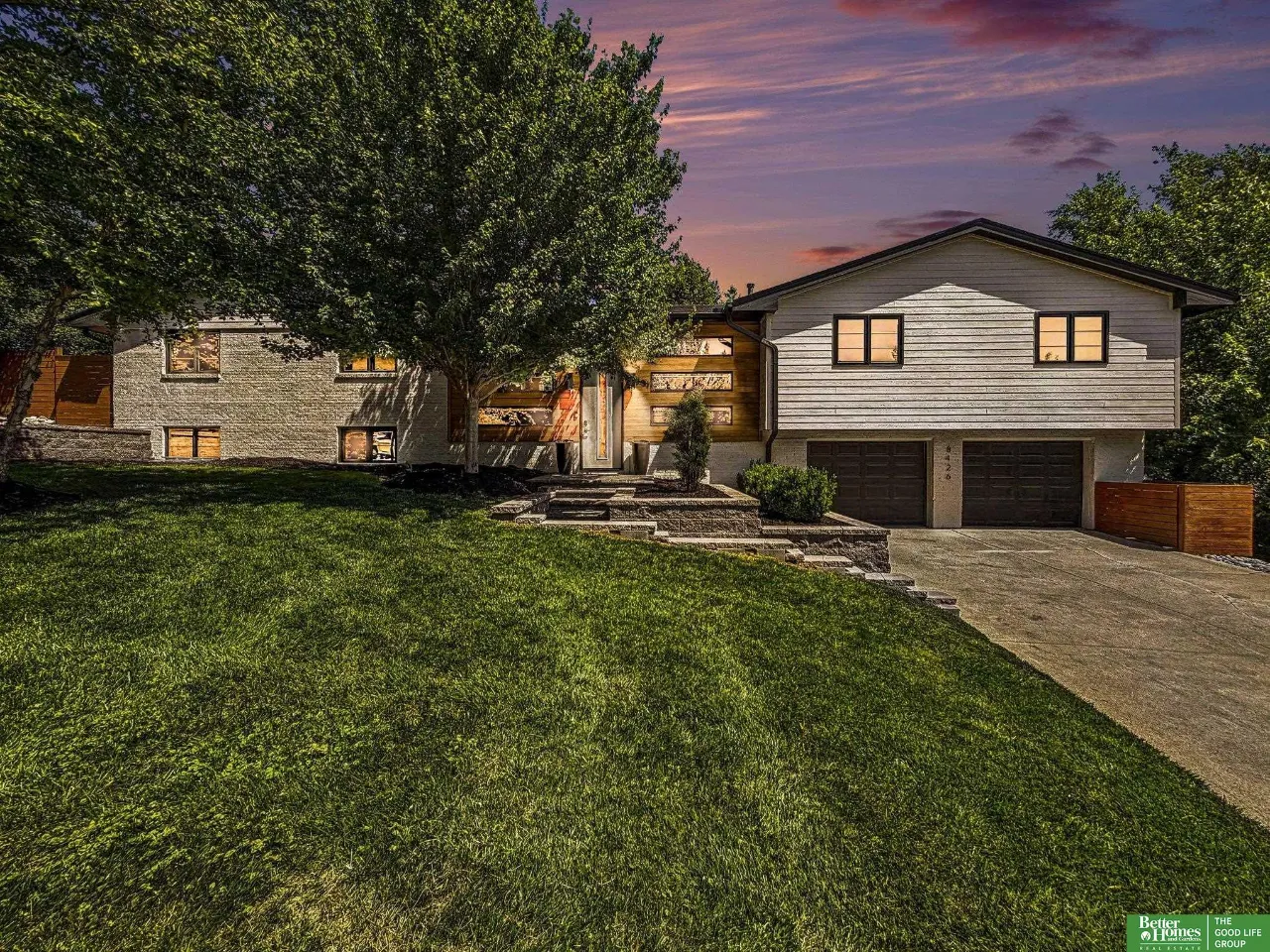 Split-level home with stone and light siding, illuminated windows, a double garage, and a broad lawn at dusk.