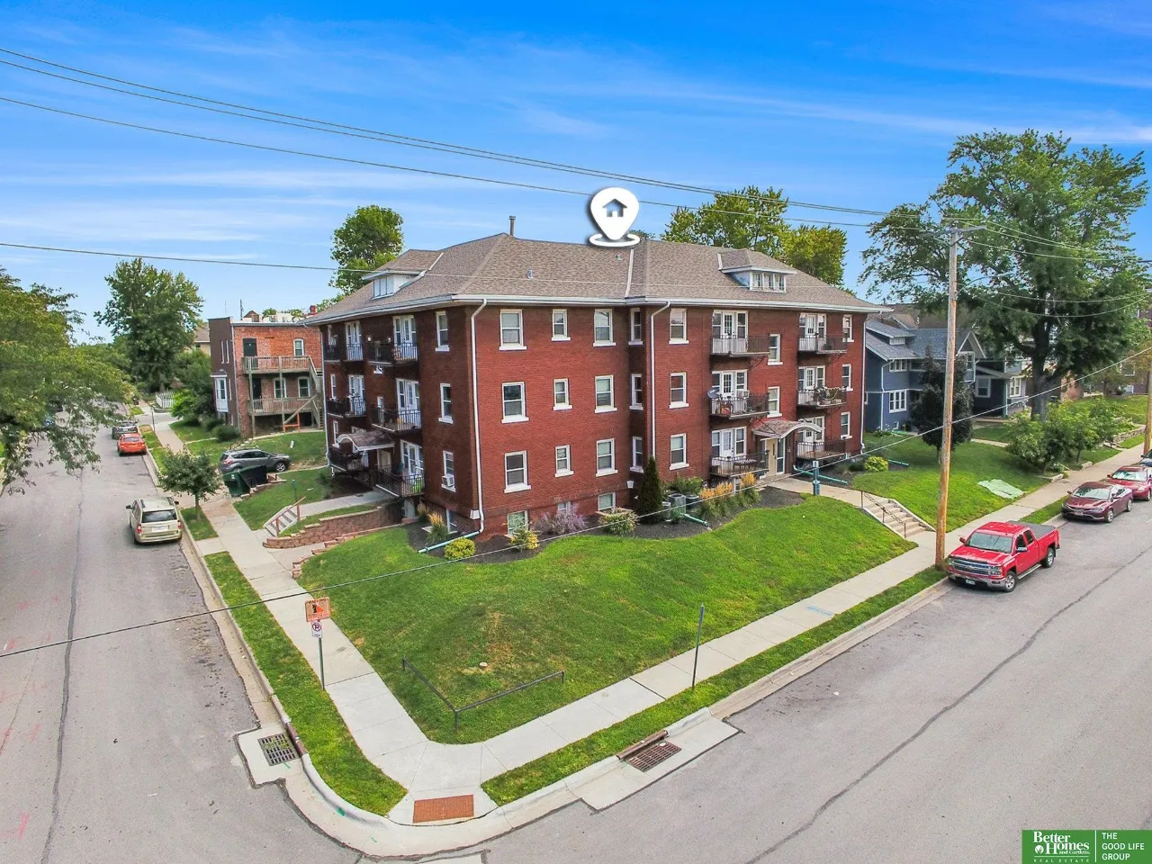 Red brick apartment building with balconies on a landscaped corner lot, viewed from above.