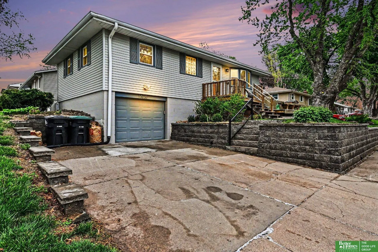 Gray split-level home with an attached garage, raised deck, and stone retaining wall at dusk.