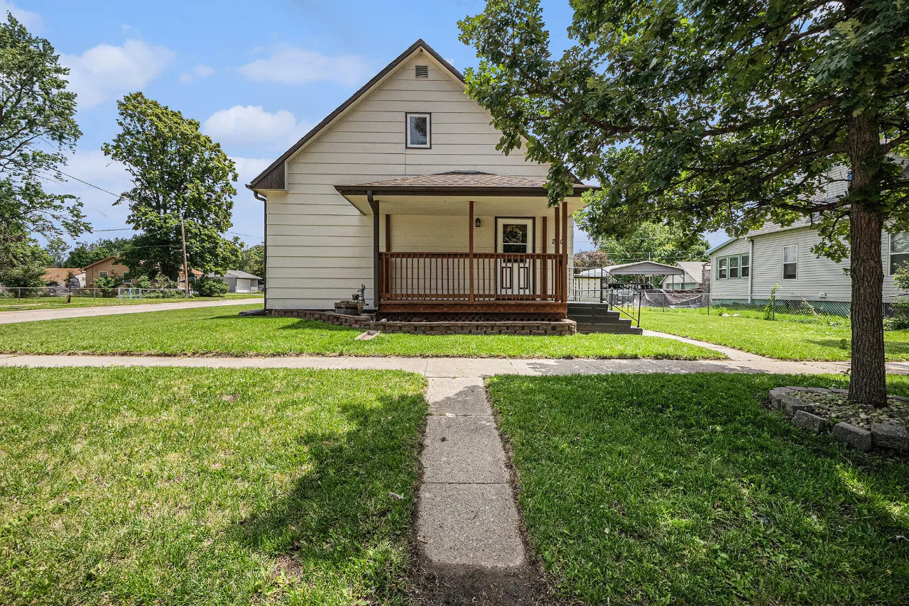 Light-colored gabled house with a covered wooden porch, front lawn, and mature shade tree.