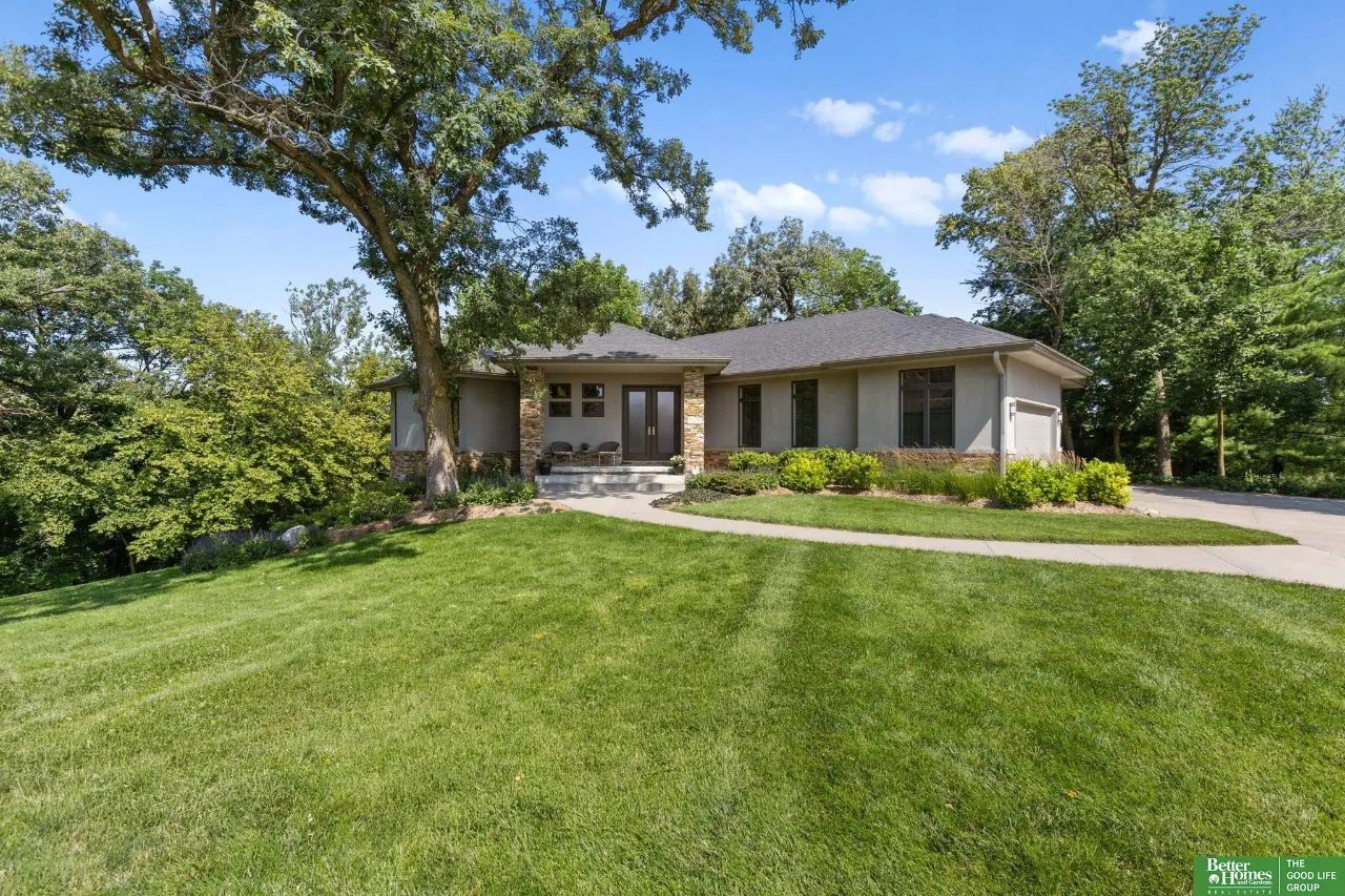 Single-story gray home with stone columns, a broad lawn, and mature trees.
