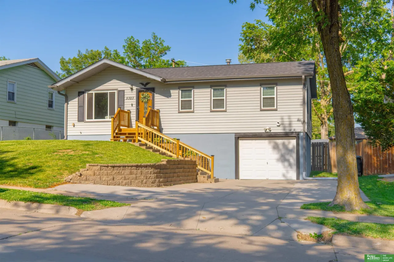 Single-story home with raised wooden entry stairs, attached garage, sloped lawn, and a large shade tree.