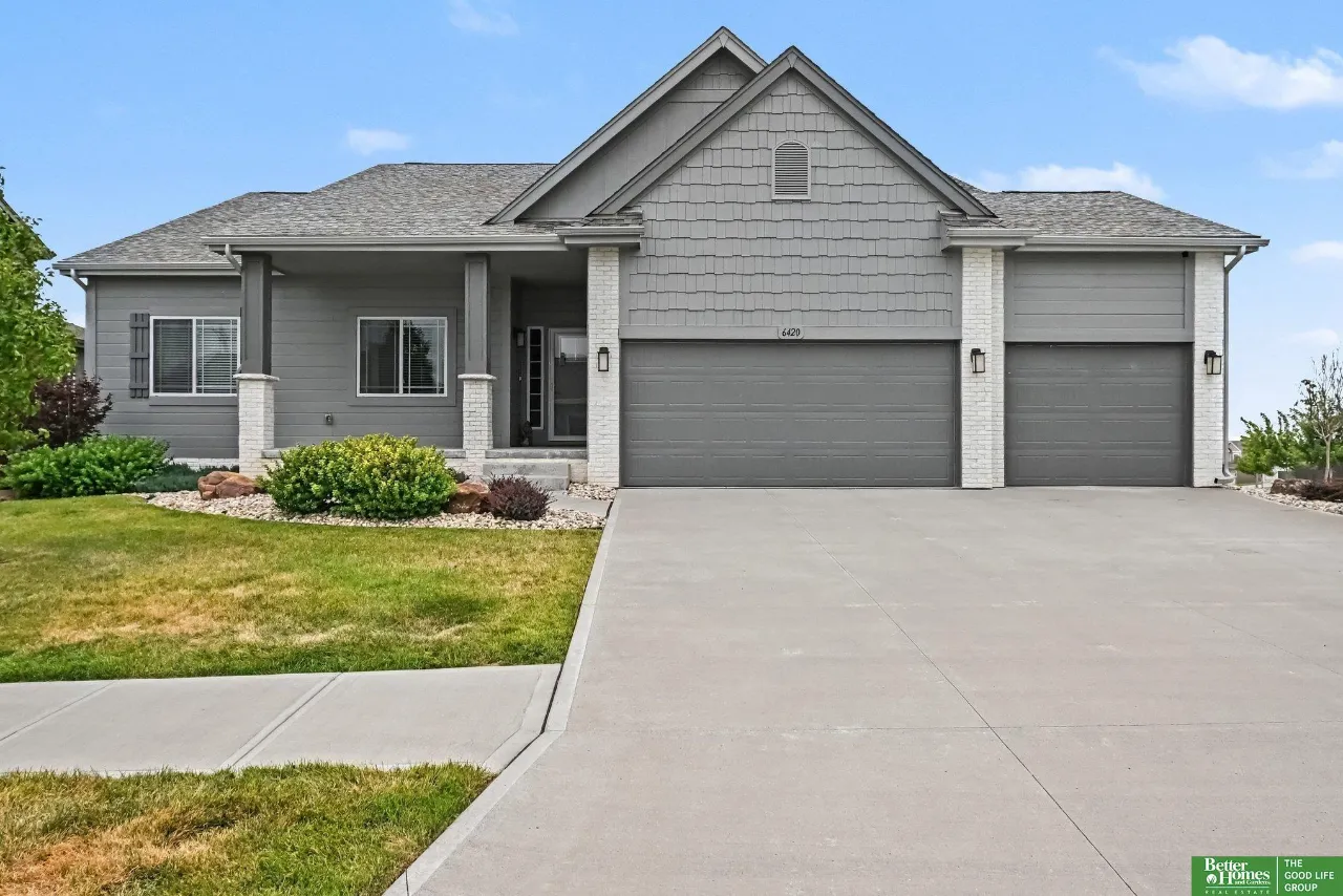 Gray gabled house with white brick accents, two garages, landscaped lawn, and a wide concrete driveway.