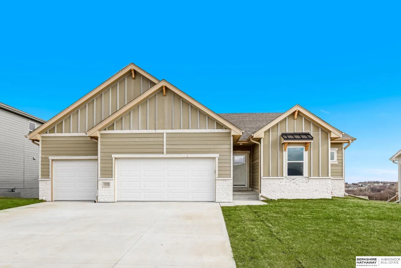 Beige gabled home with two garage doors, white stone accents, a wide driveway, and green lawn.