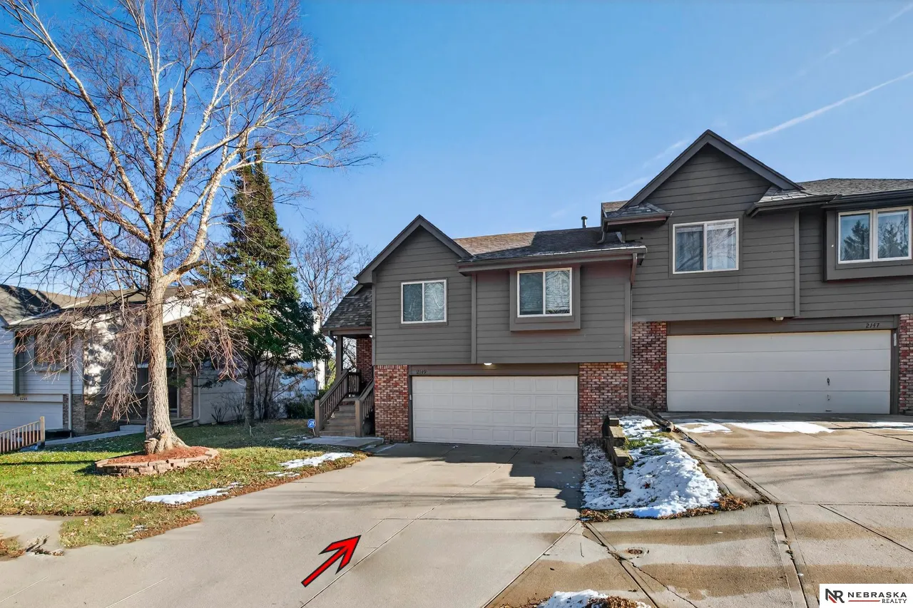 Gray townhouse with brick lower walls, white garage doors, and a red arrow pointing toward the driveway.