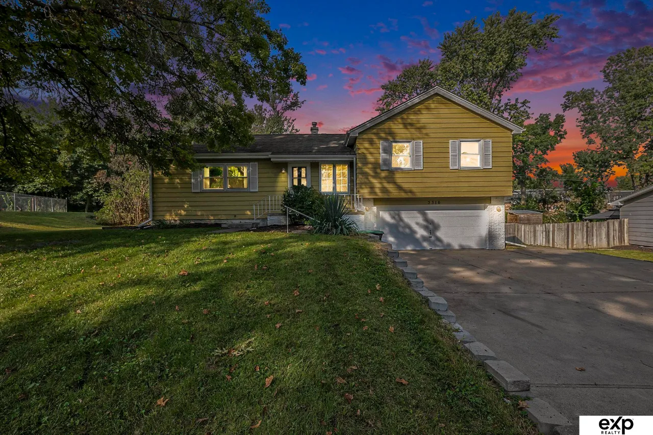 Green split-level house with a two-car garage and broad lawn beneath a colorful dusk sky.