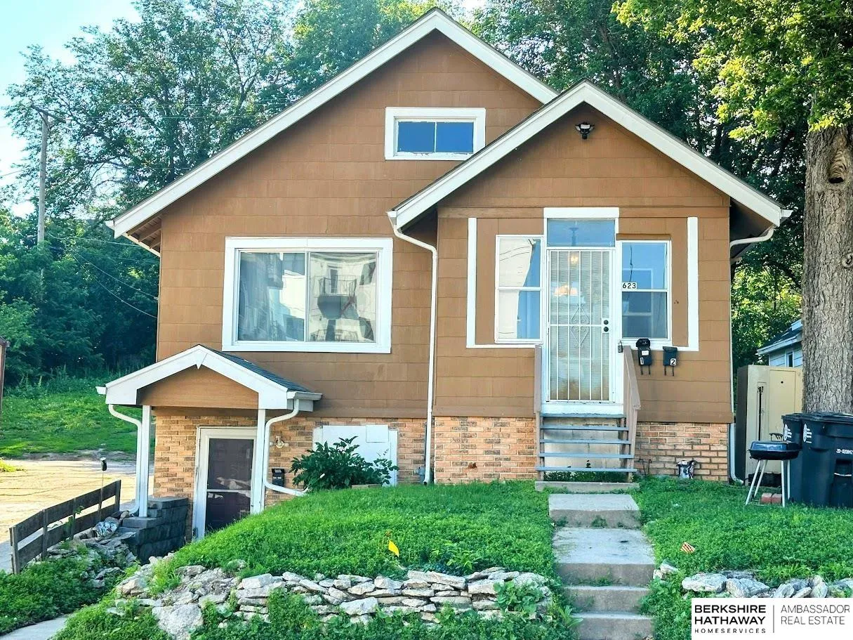 Brown two-story house with a brick lower level, white-trimmed entrance, and stone landscaping along the front path.