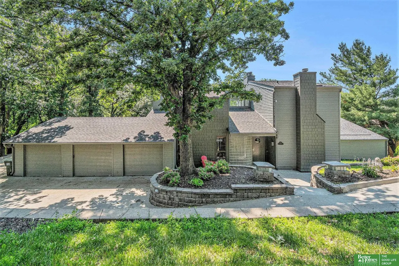 Gray multi-level house with a three-car garage, mature shade tree, and landscaped stone retaining walls.