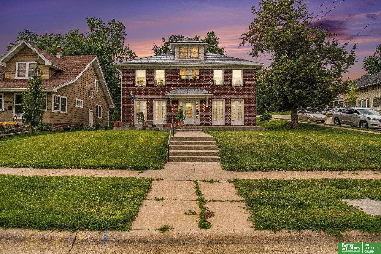 Red-brick two-story home with lit windows, front steps, and a broad lawn at dusk.