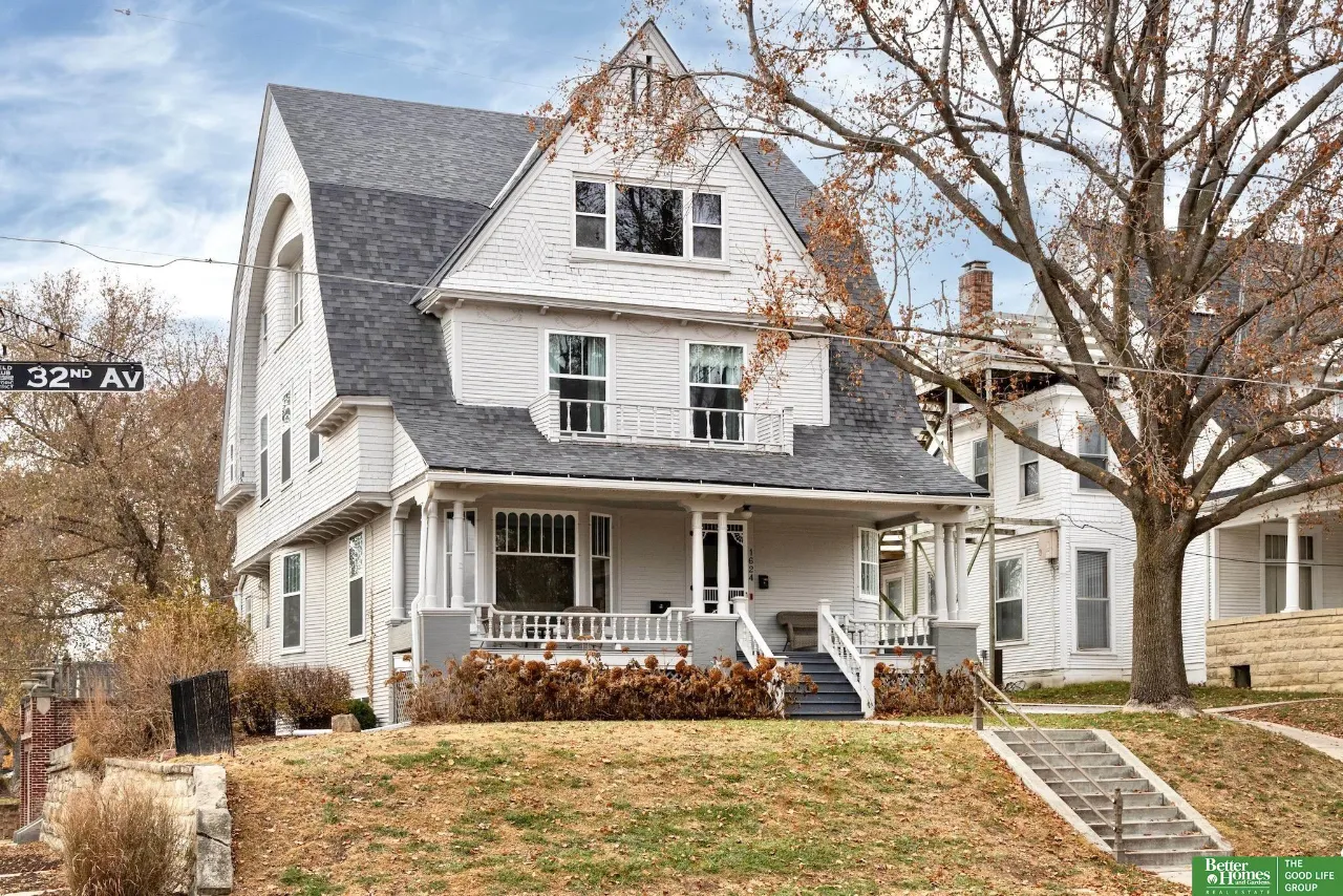 White multi-story house with a columned porch, steep roofs, and front steps on a leafy autumn street.