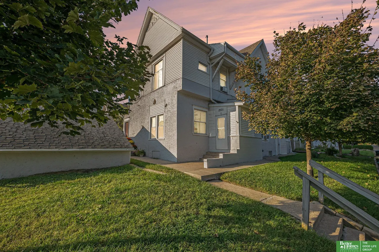 Gray multi-story house with a front porch and steps, framed by mature trees at sunset.